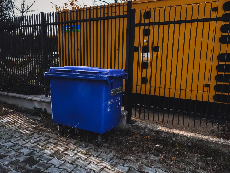 A blue trash can sitting next to a fence.