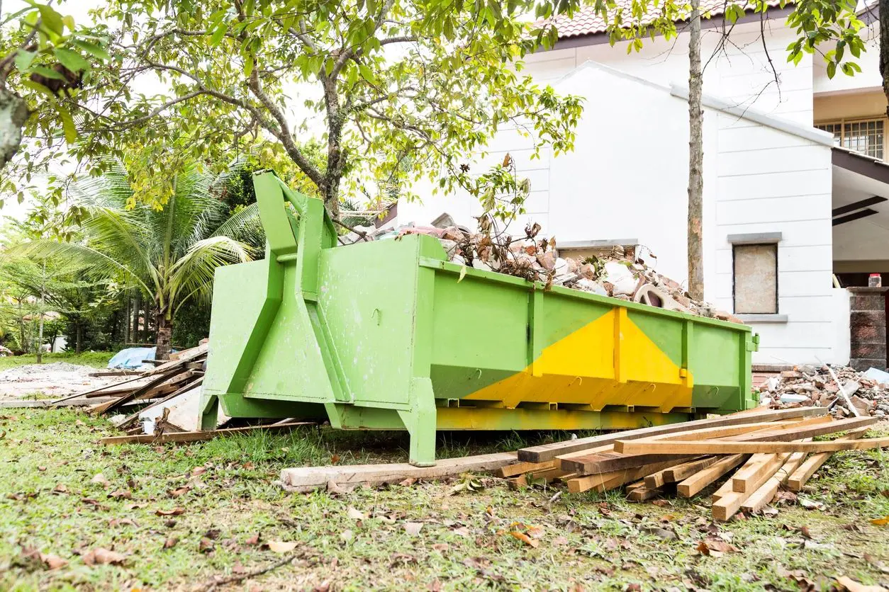 A green dumpster with wood piled up in front of it.